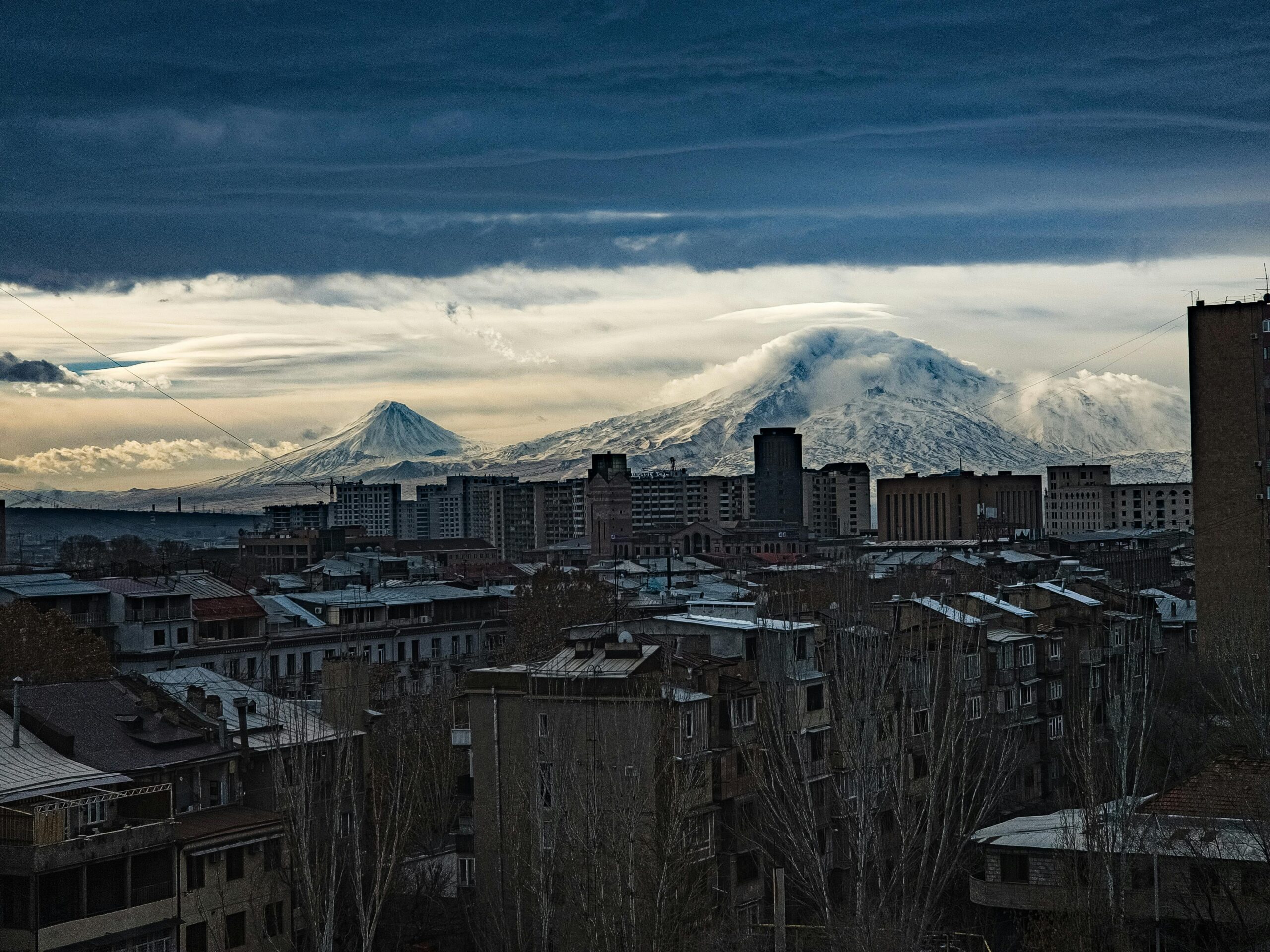 A breathtaking view of a city skyline with snow-covered mountains under a dramatic cloudy sky.